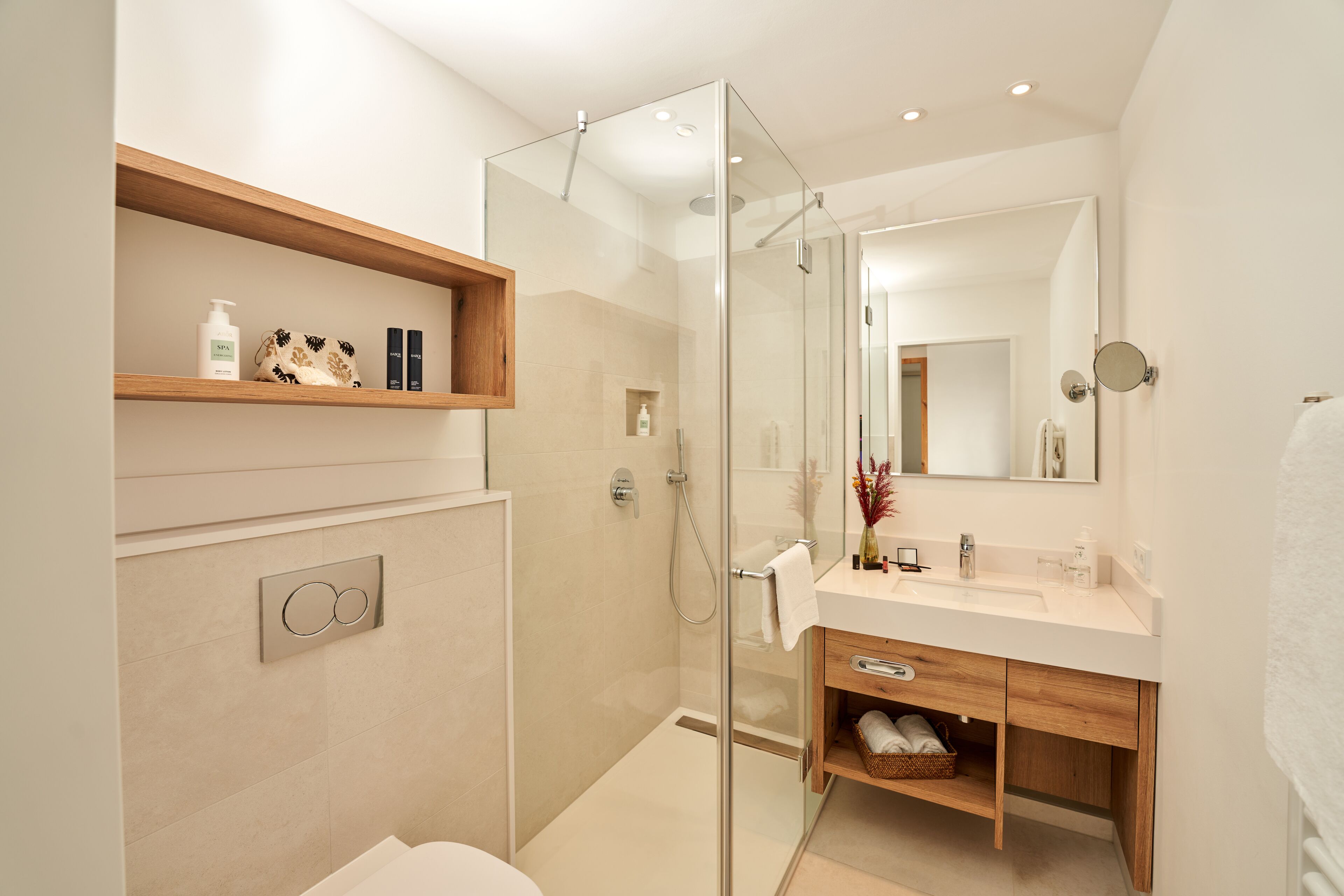 A modern bathroom with a glass-enclosed shower, a wooden shelf with toiletries, a white sink, and a mirror.