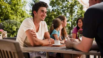 Un groupe de personnes est assis à une table à l’extérieur, recouverte d’une nappe blanche, et est entouré de verdure.