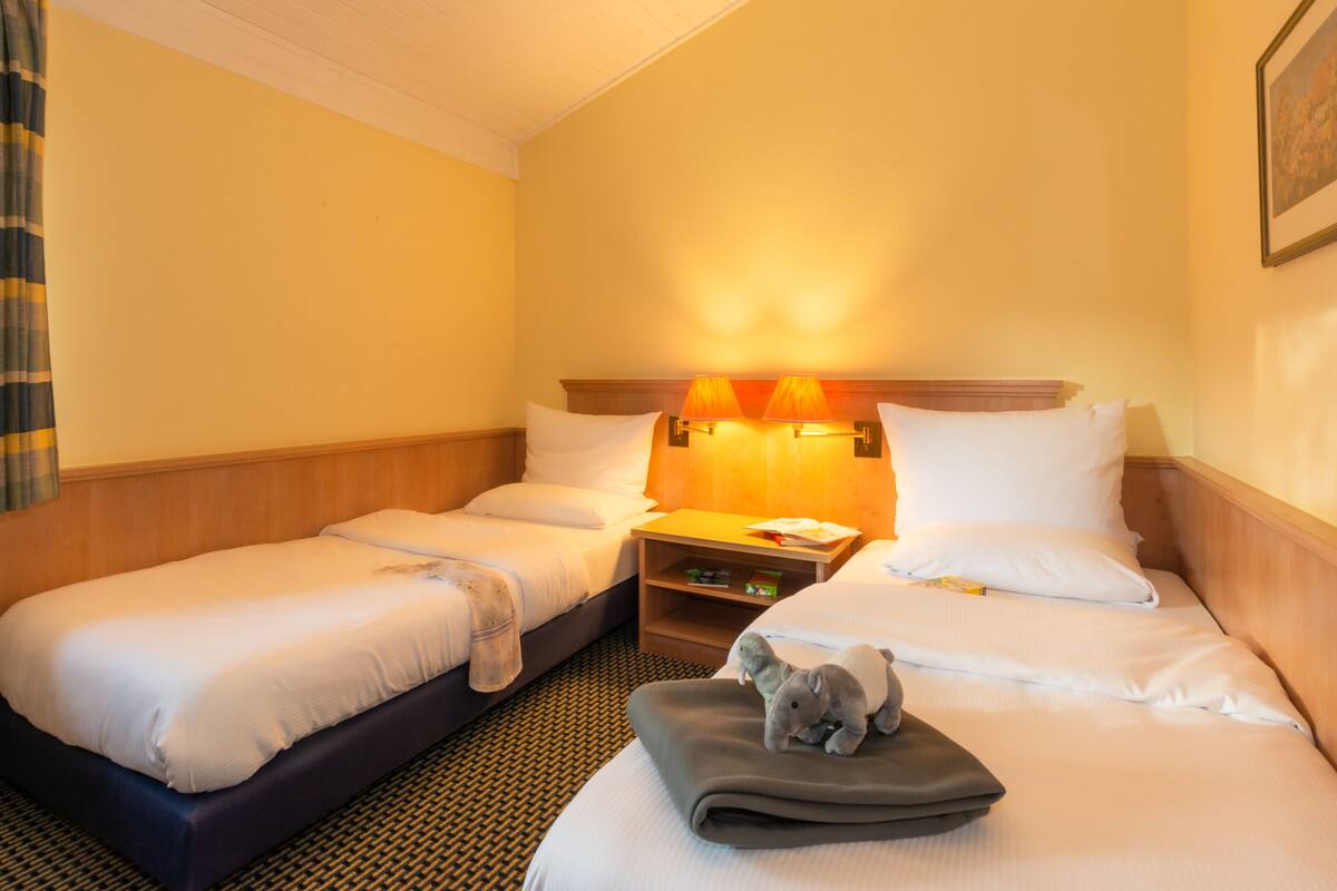 A hotel room with two single beds, a nightstand with a lamp, and a plush toy on the bed.