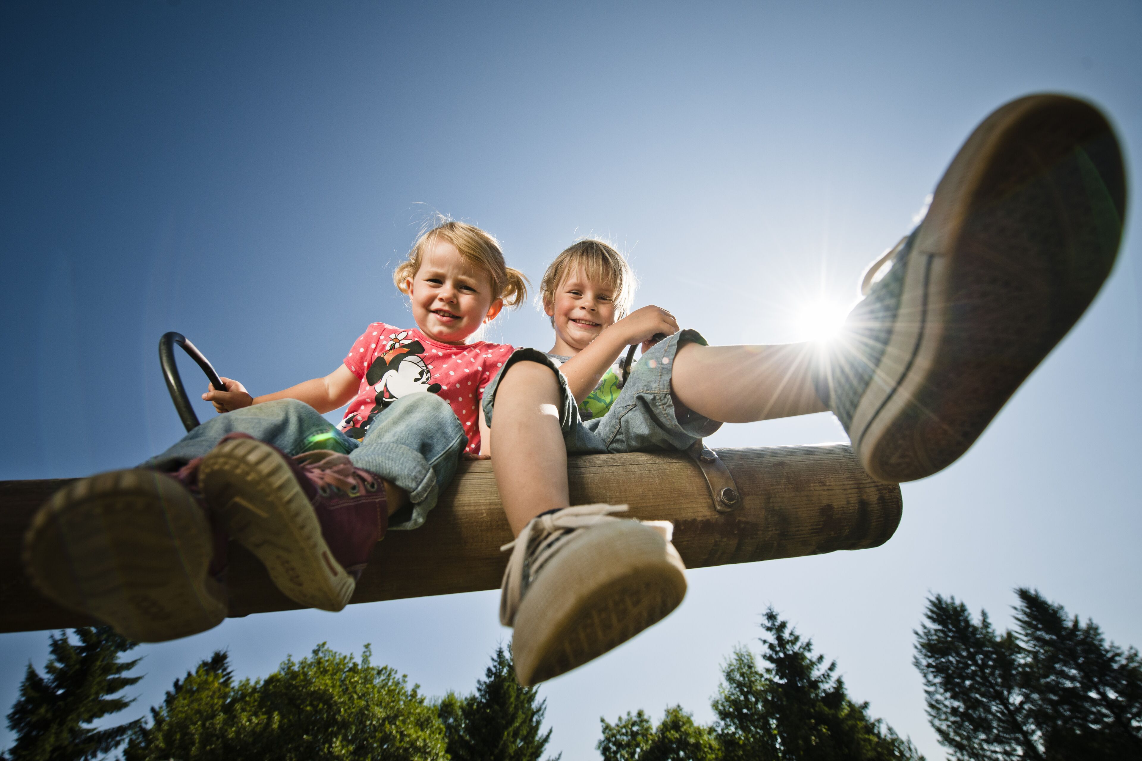 Two children sitting on a wooden swing with a clear sky in the background.