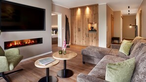 A modern living room with a flat-screen TV, a fireplace, a grey sofa, and a round wooden table. - Sporthotel Grafenwald (Daun)