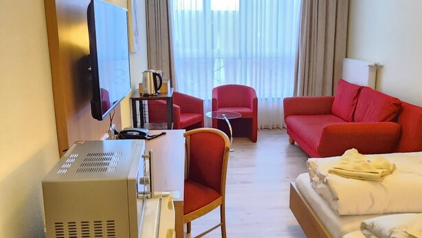 A hotel room with a bed, a desk with a computer, a chair, a red sofa, and a television. - Posthotel Traube (Donauwoerth)