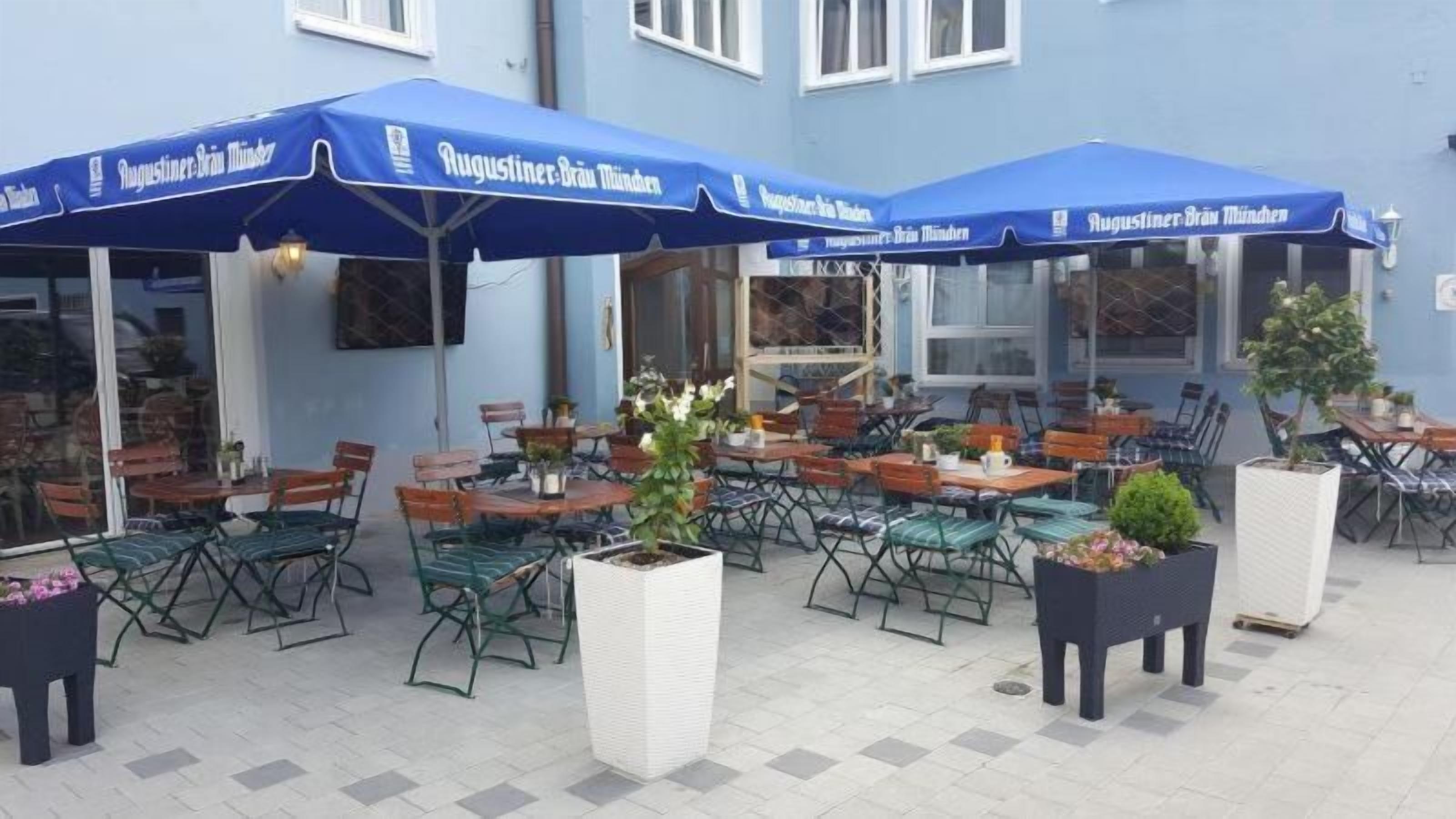 Un espacio al aire libre con mesas y sillas, cubierto por sombrillas azules, y plantas en macetas.