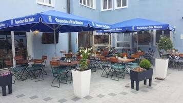 Ein Sitzbereich im Freien mit Tischen und Stühlen, beschattet von blauen Sonnenschirmen, mit Pflanzkübeln.