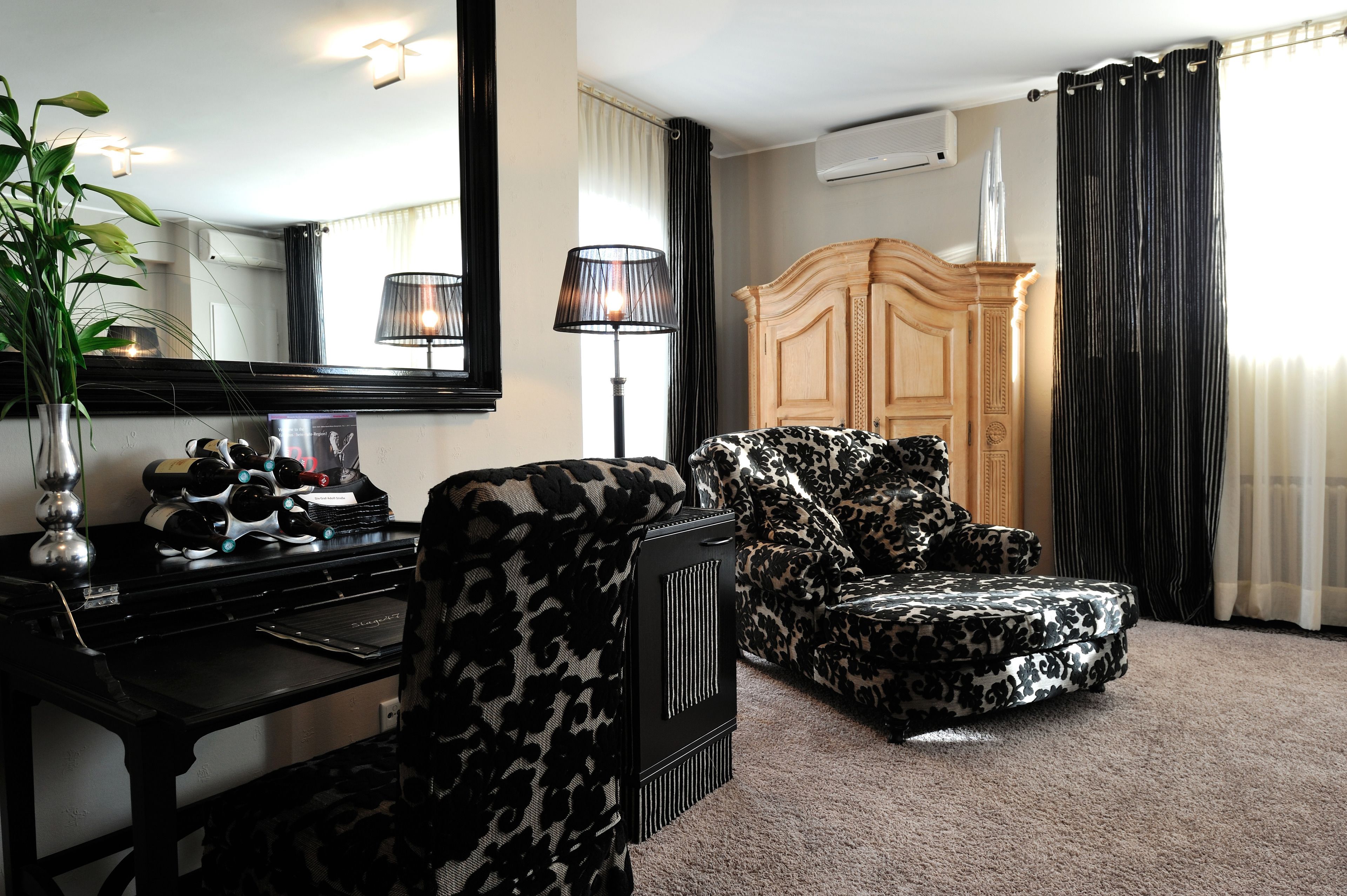 A room with a black and white patterned armchair, a wooden cabinet, a desk with a mirror, and a potted plant.