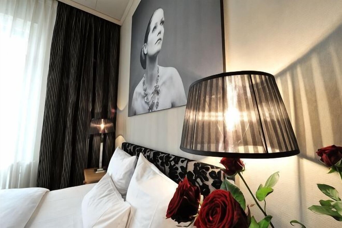 Ein Hotelzimmer mit einem Bett, einer Lampe, einem gerahmten Porträt und einer Vase mit roten Rosen.