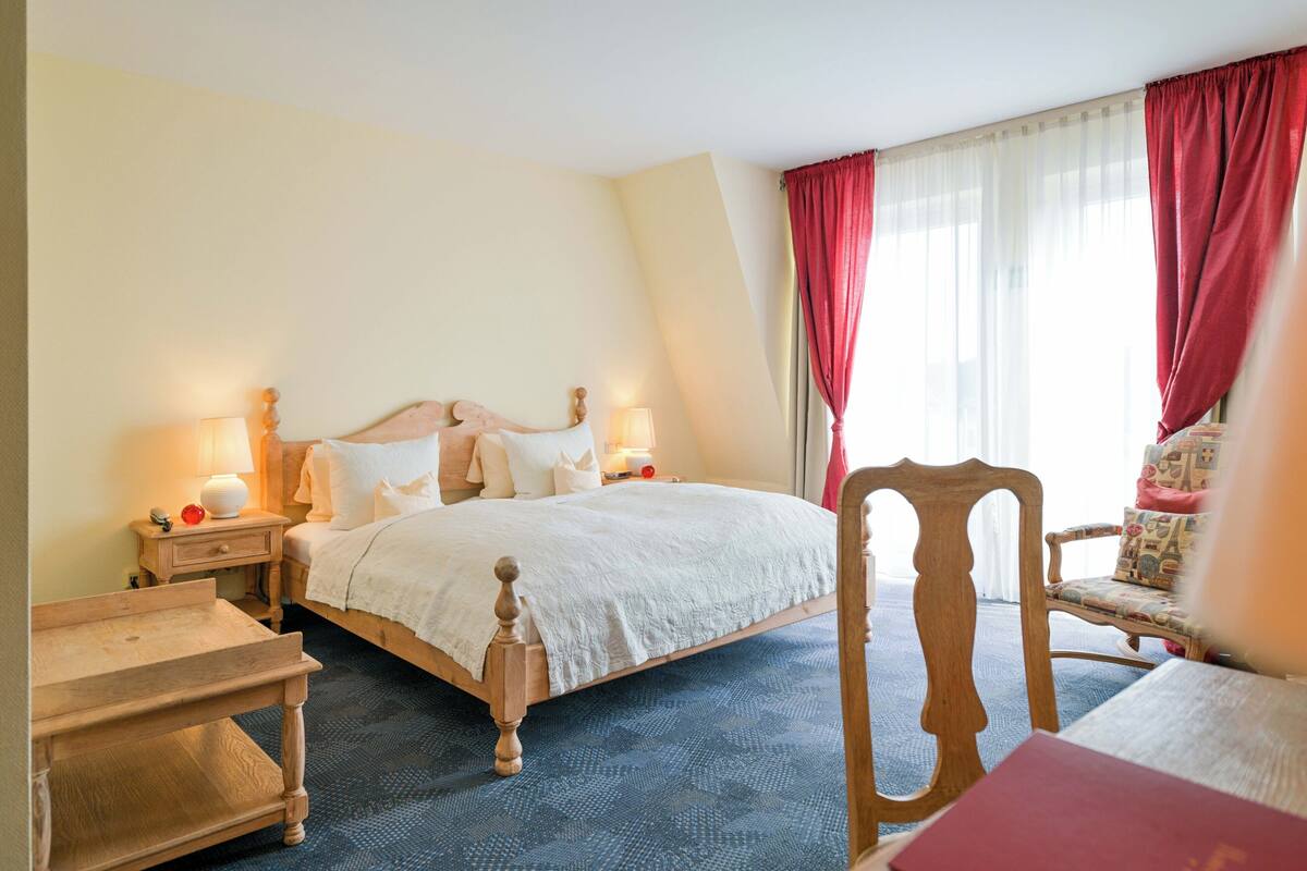 Una habitación de hotel con cama, mesitas de noche, una silla y un ventanal con cortinas.
