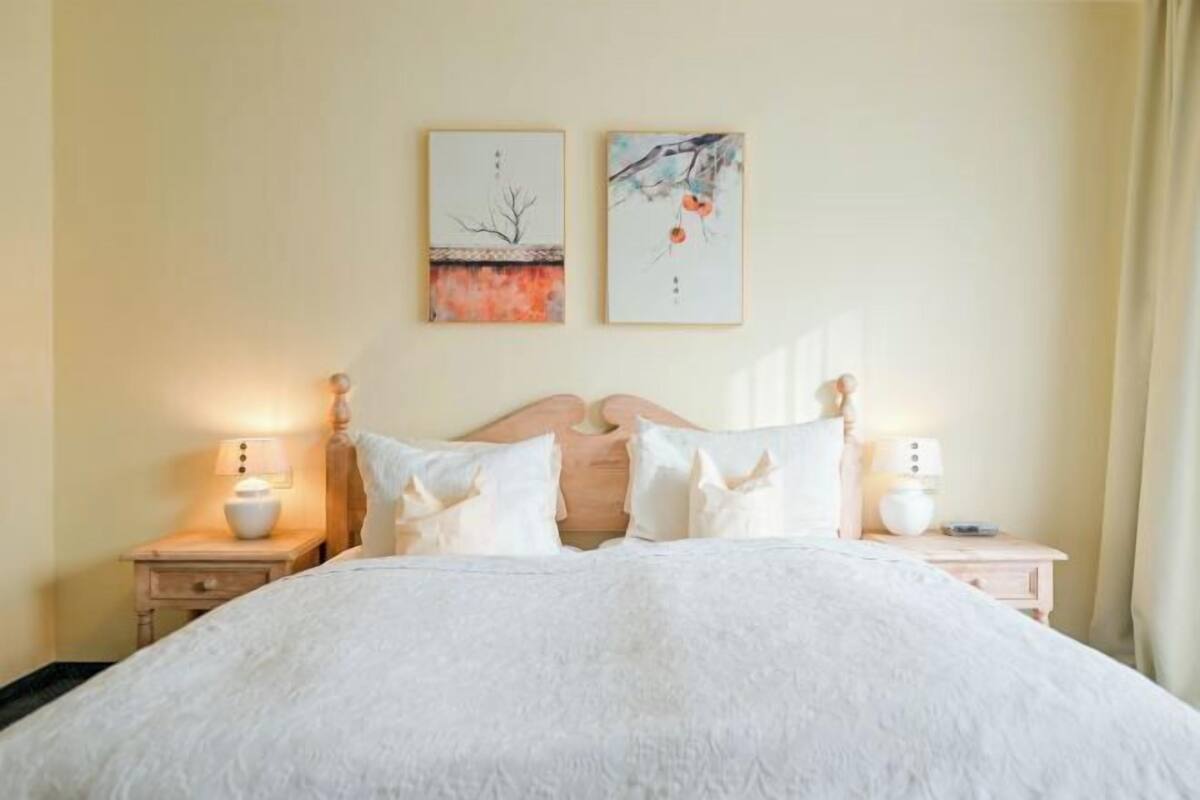 Una cama bien tendida con ropa de cama blanca, dos mesitas de noche con l ámparas y dos obras de arte colgadas en la pared.