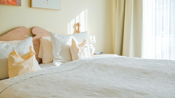 A neatly made bed with a white duvet and pillows, a wooden headboard, a bedside table with a lamp, and a window with curtains. - Hotel Lessing (Düsseldorf)