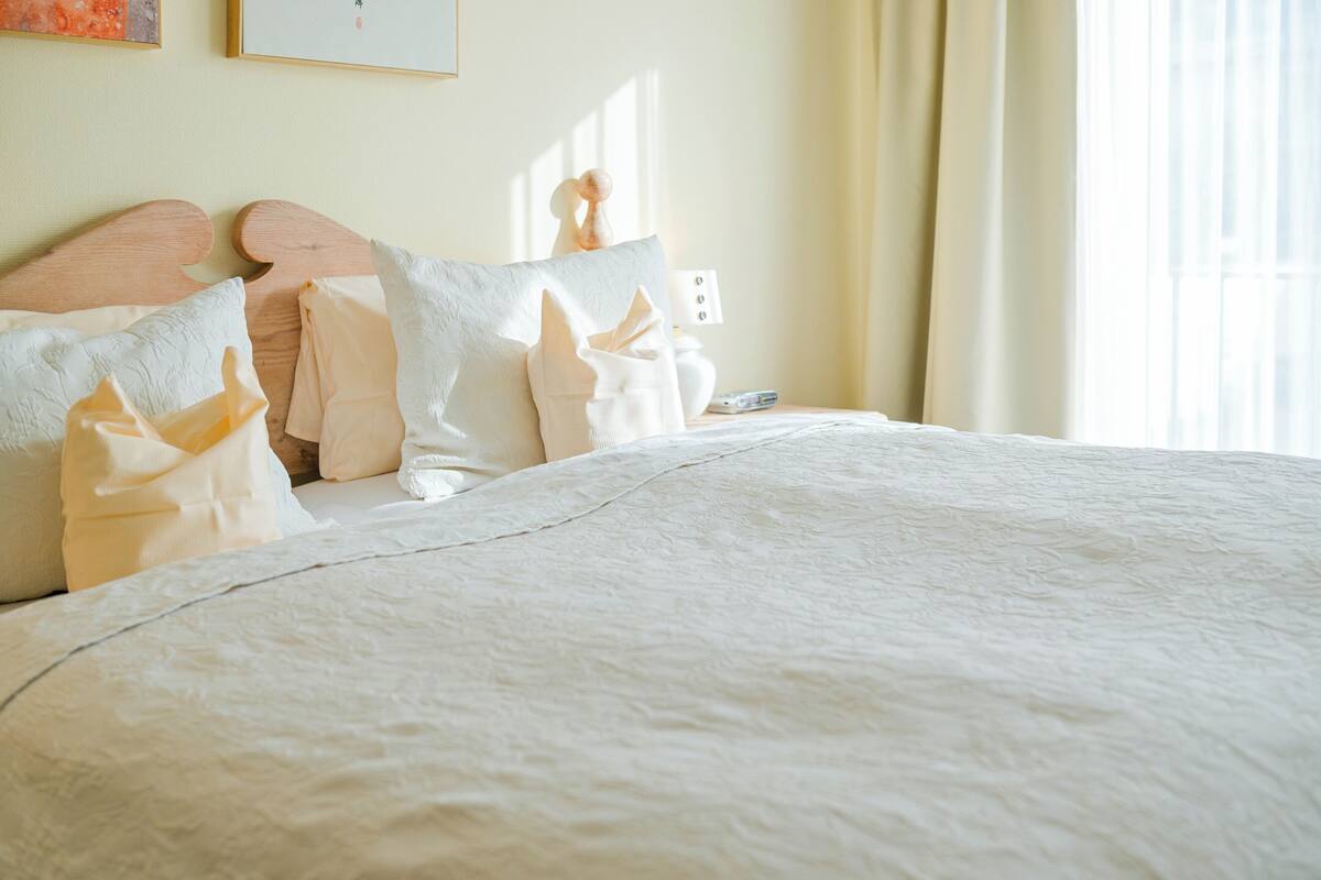 Una cama bien hecha con edredón blanco y almohadas, cabecera de madera, mesita de noche con lámpara y ventana con cortinas.