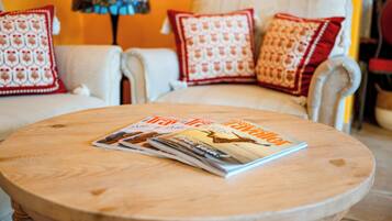 Una mesa redonda de madera con una revista, un sillón blanco con cojines estampados en rojo, y una lámpara al fondo.