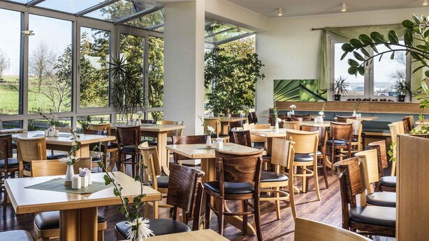 Ein geräumiges Restaurant mit hohen Decken, großen Fenstern und Holztischen und -stühlen.