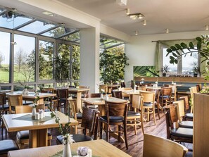 Een ruime restaurantruimte met hoge plafonds, grote ramen en houten tafels en stoelen.