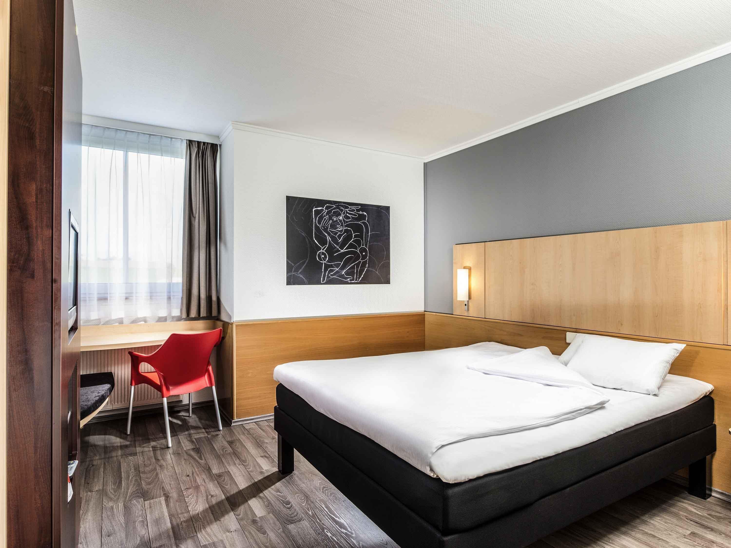 A hotel room with a bed, a desk, a chair, a wall art piece, and a light fixture.