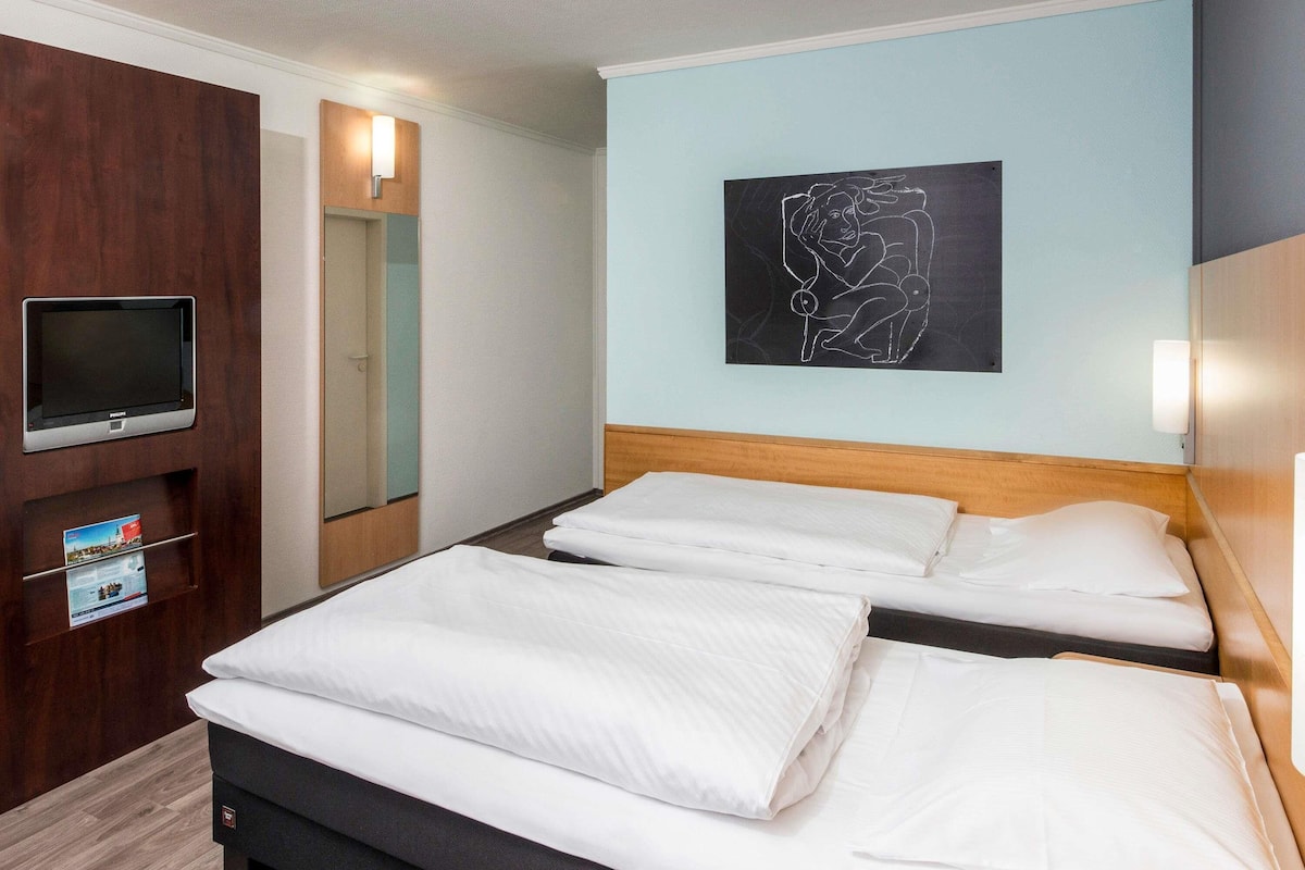 Une chambre d’hôtel avec un lit, un téléviseur, une œuvre d’art accrochée au mur et un panneau en bois sur le côté gauche.