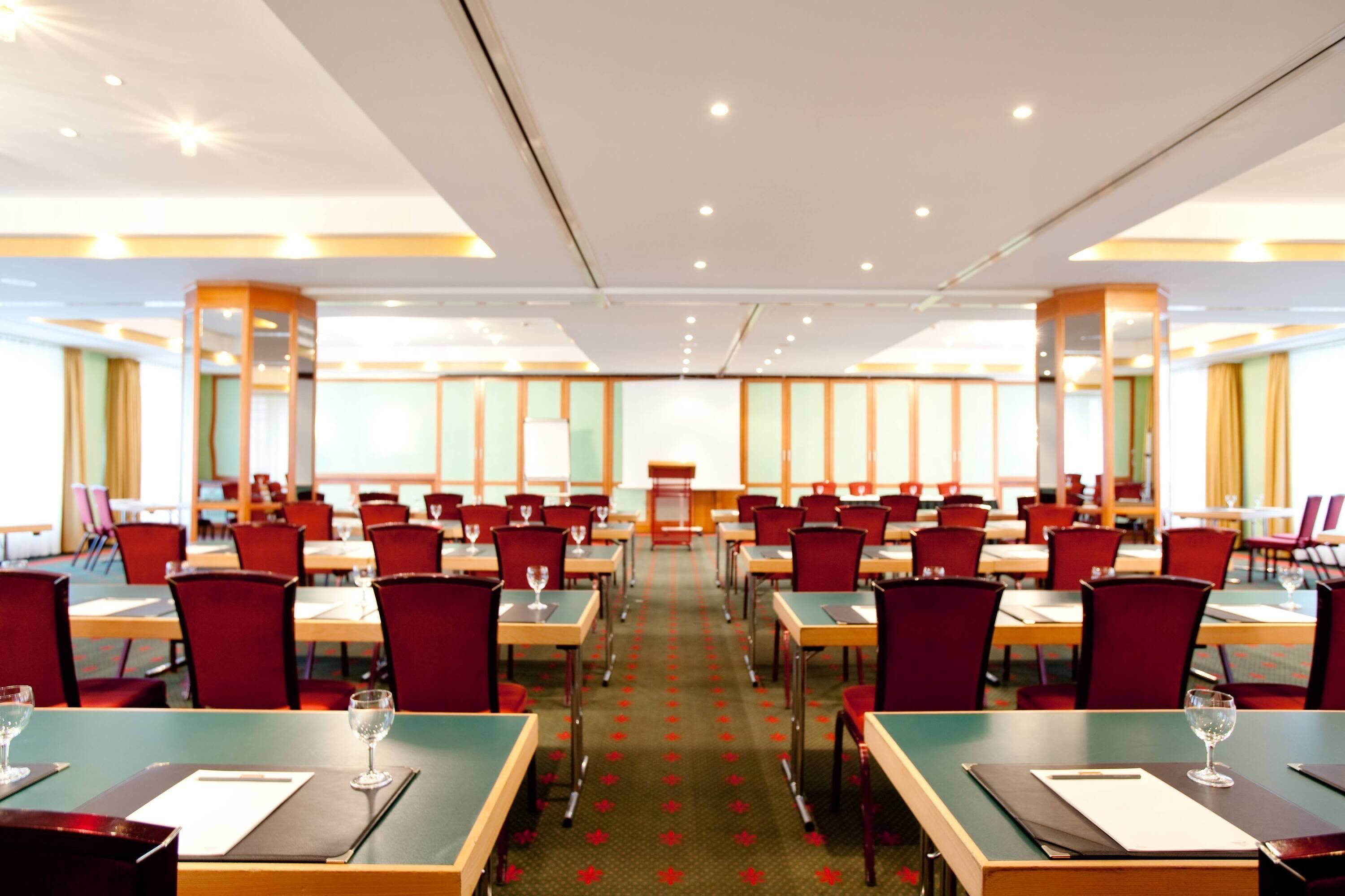 Comedor de hotel con varias mesas y sillas, suelo alfombrado y un ventanal grande al fondo.