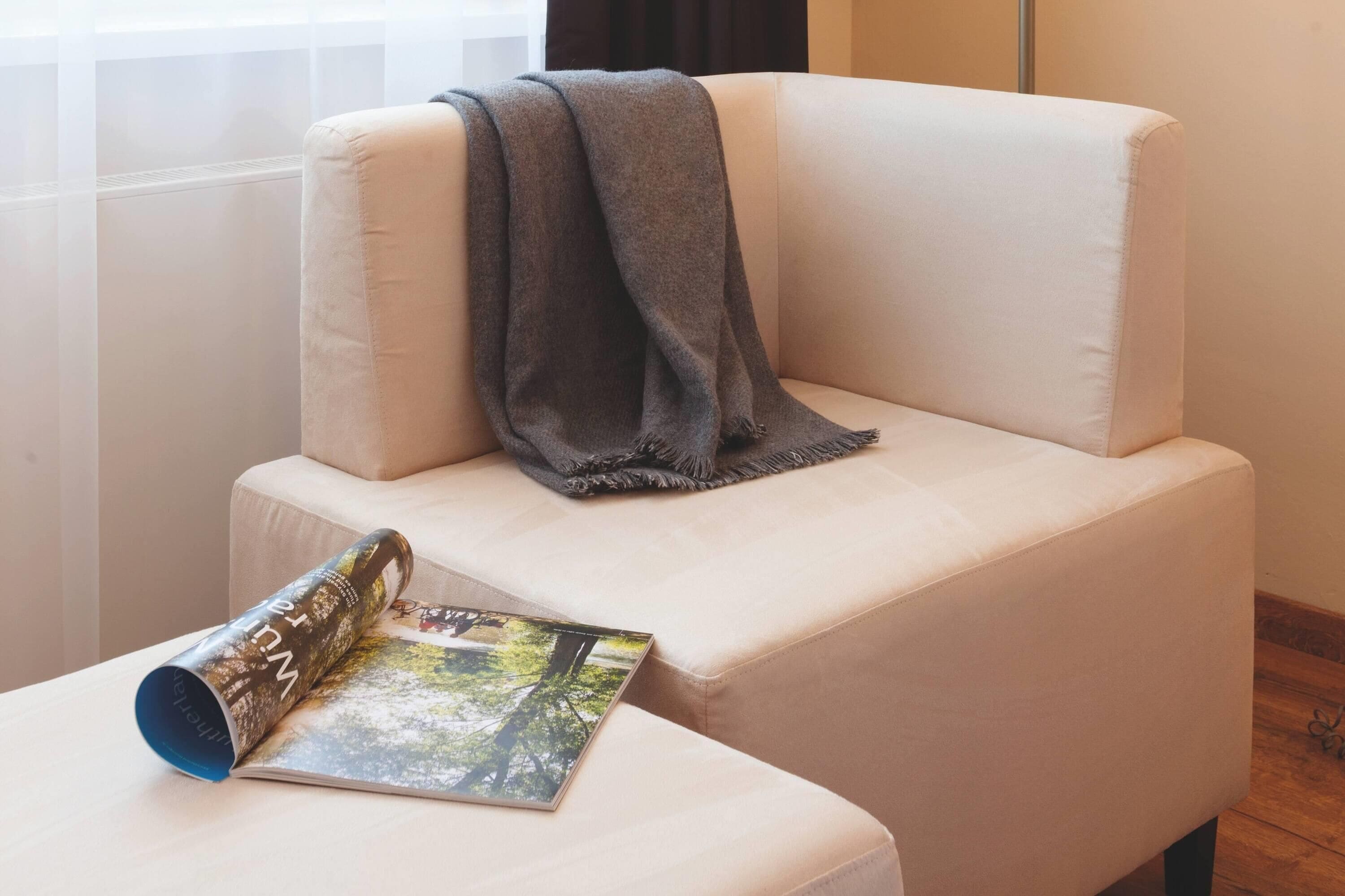 Una silla blanca acolchada con una manta gris, una revista enrollada y una ventana con cortinas traslúcidas.