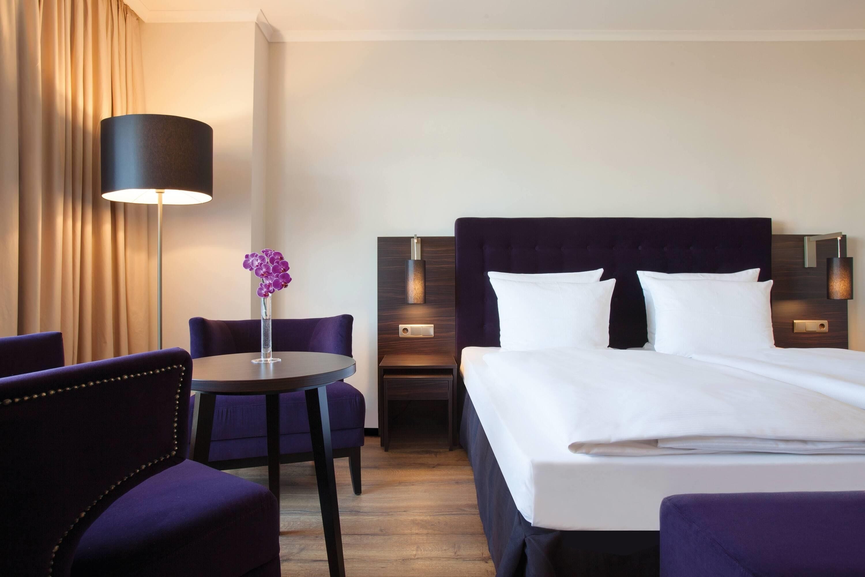Ein Hotelzimmer mit einem Bett, zwei Nachttischen, einer Lampe, einem Sessel, einem kleinen Tisch und einer Vase mit Blumen.