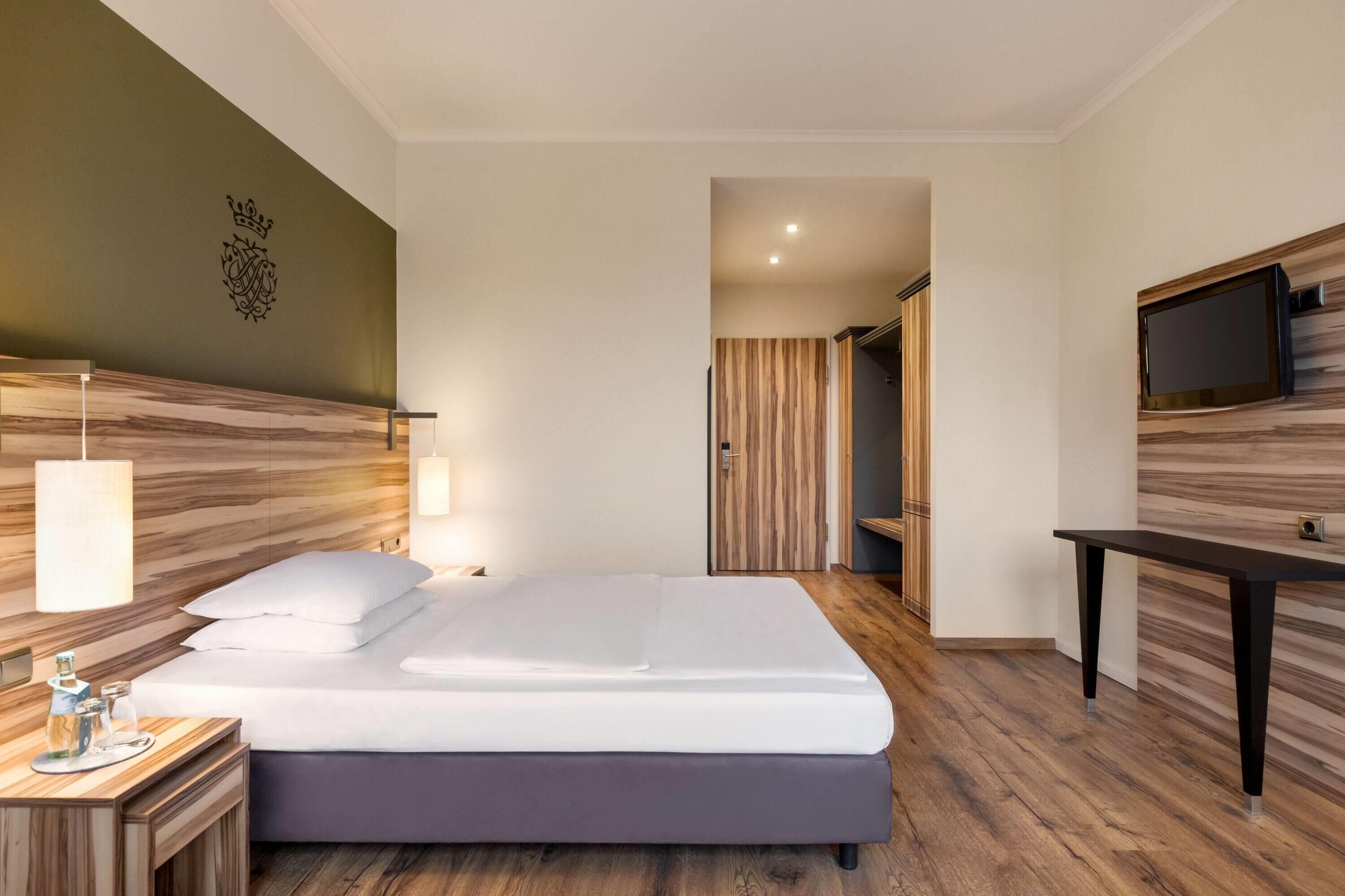 A modern hotel room with a large bed, bedside table, lamp, and a flat-screen TV mounted on the wall.