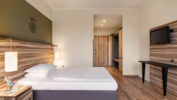A modern hotel room with a large bed, bedside table, lamp, and a flat-screen TV mounted on the wall.