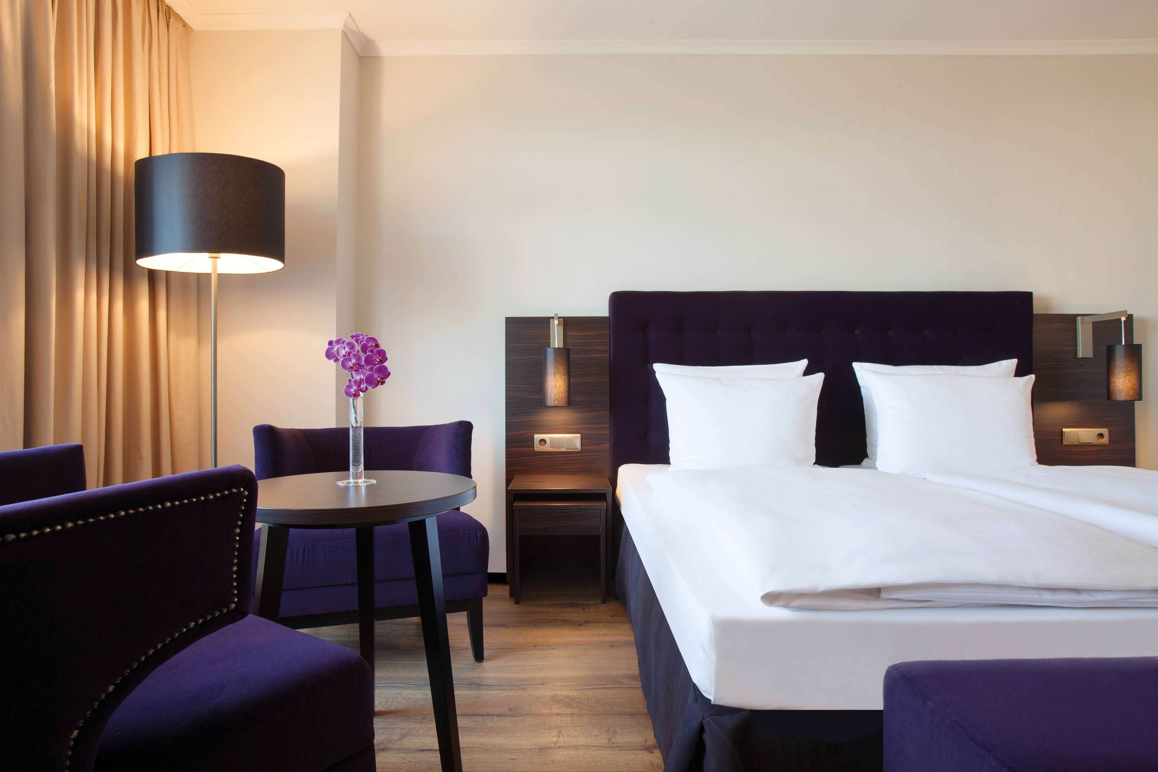A hotel room with a large bed, a small table with a chair, a lamp, and a vase with flowers.