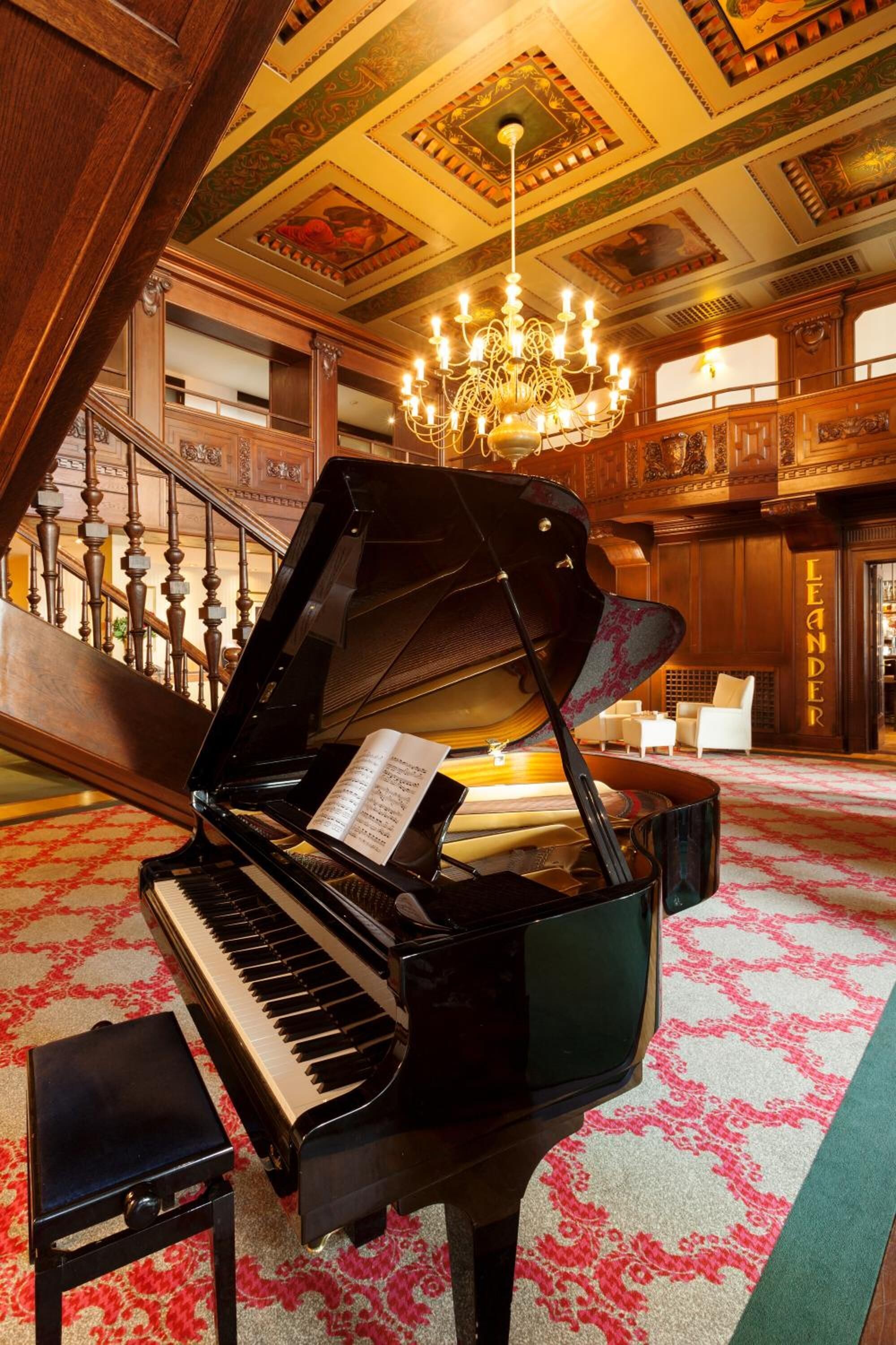 A grand piano in a spacious lobby with ornate wood paneling, a chandelier, and a patterned carpet.