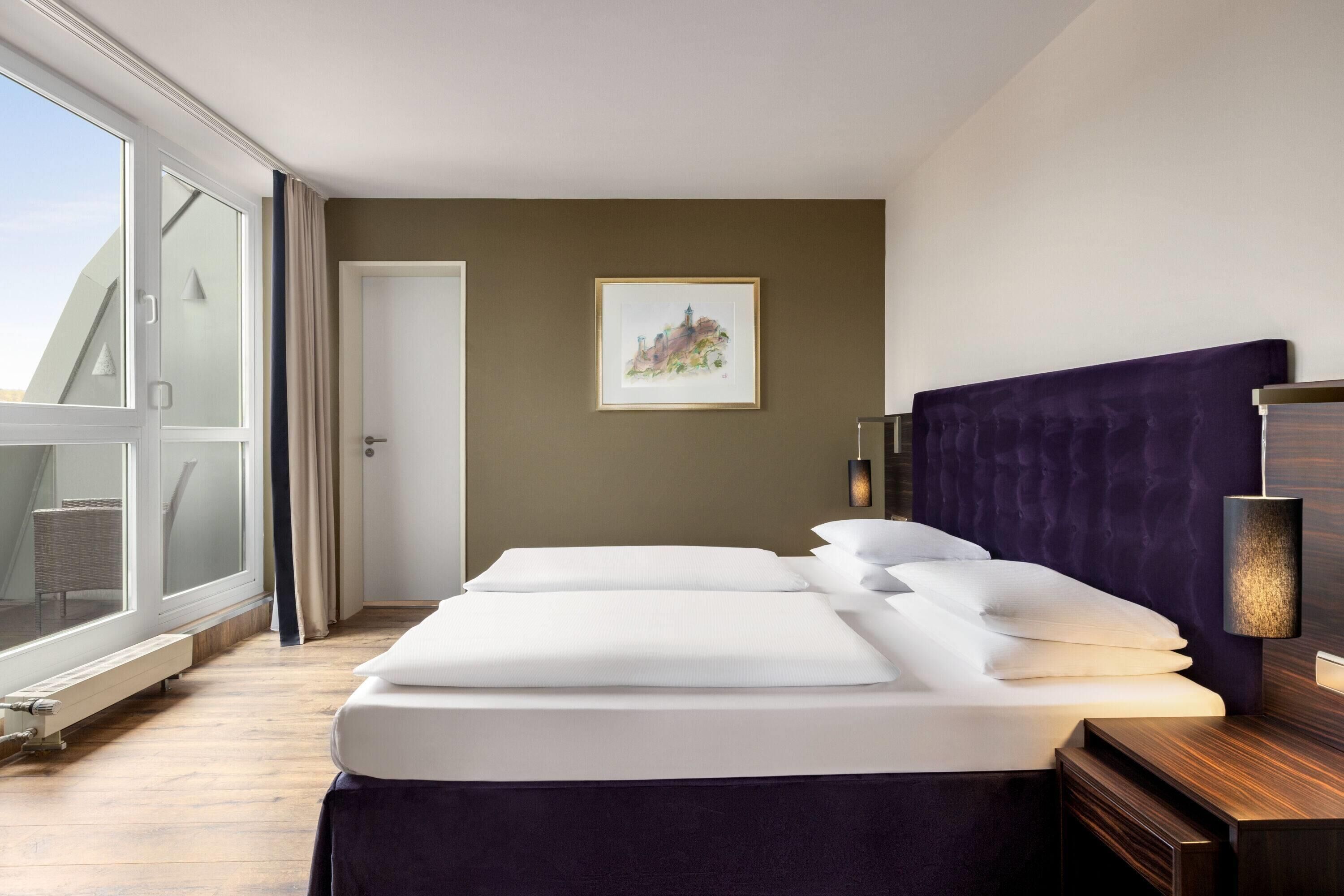 Habitación de hotel con cama, mesita de noche, lámpara de pared, puerta y ventana con vistas.
