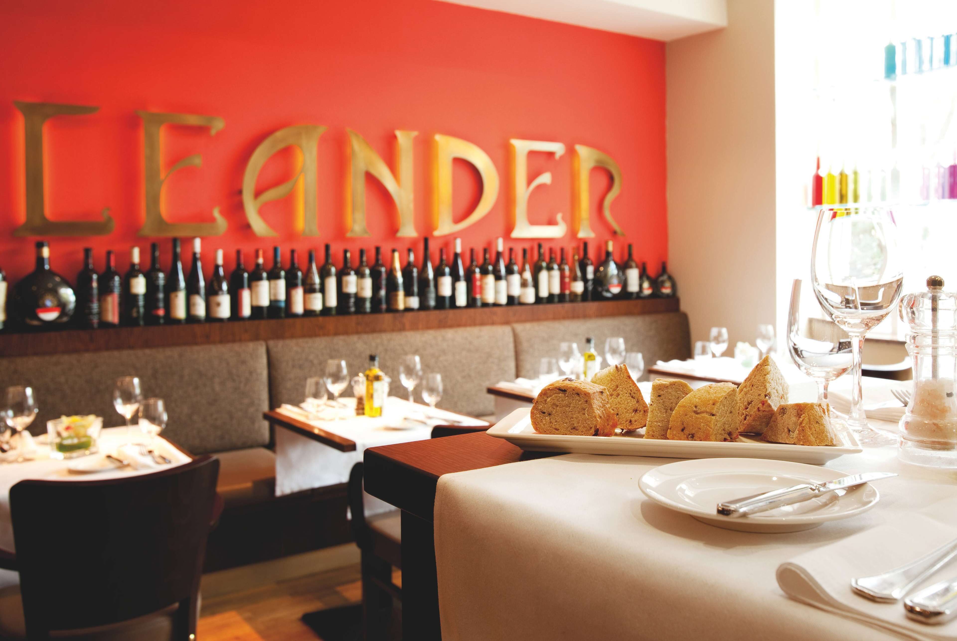 Interior de restaurante con una pared de botellas de vino, una mesa dispuesta con pan y copas de vino, y un letrero que dice "LEANDER".