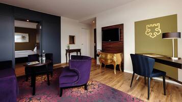 Una habitación de hotel con una cama, un escritorio, una silla, un sillón púrpura, una mesita de noche con una lámpara y un televisor en la pared.