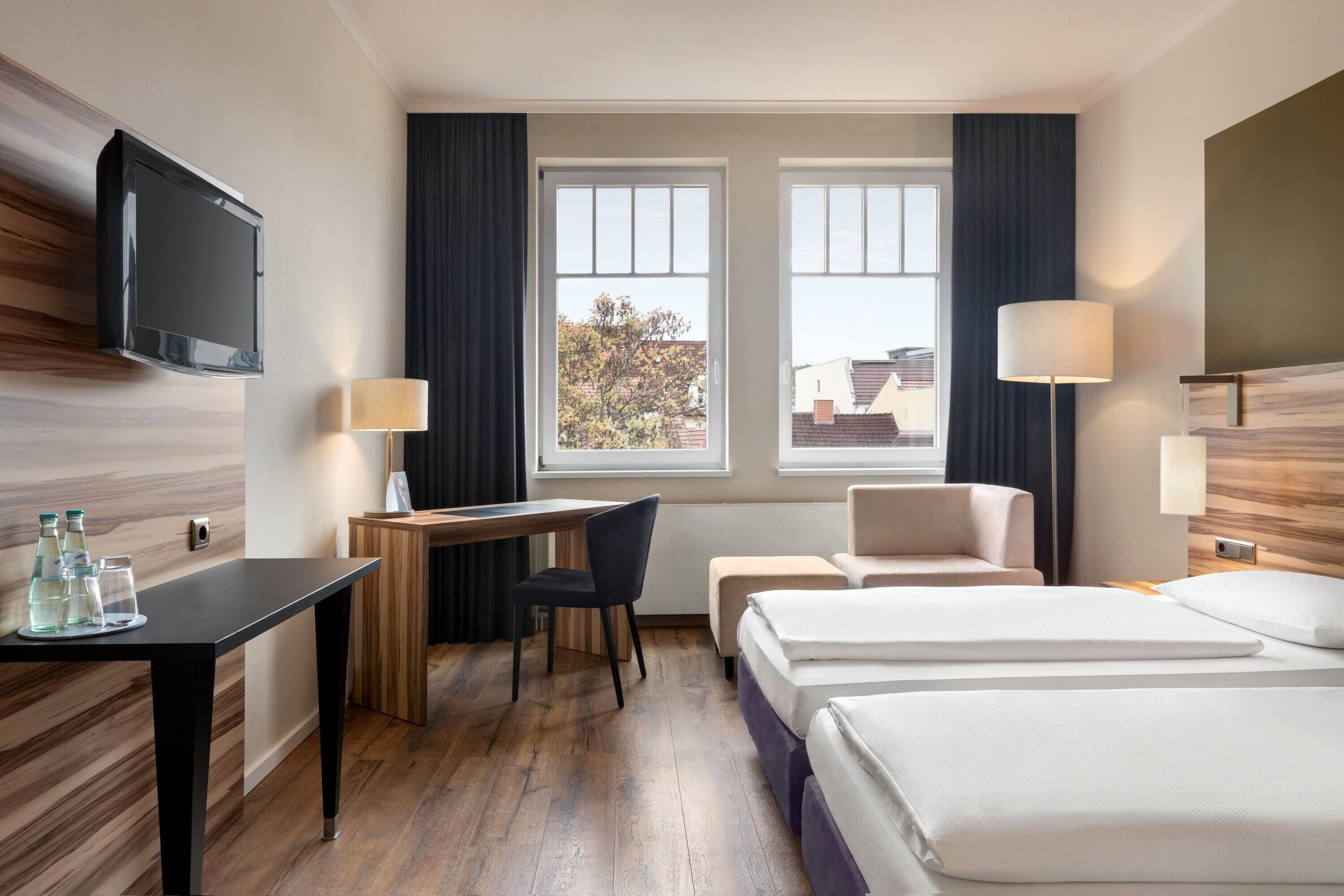 Habitación de hotel con escritorio, dos camas, televisor y ventana con cortinas.