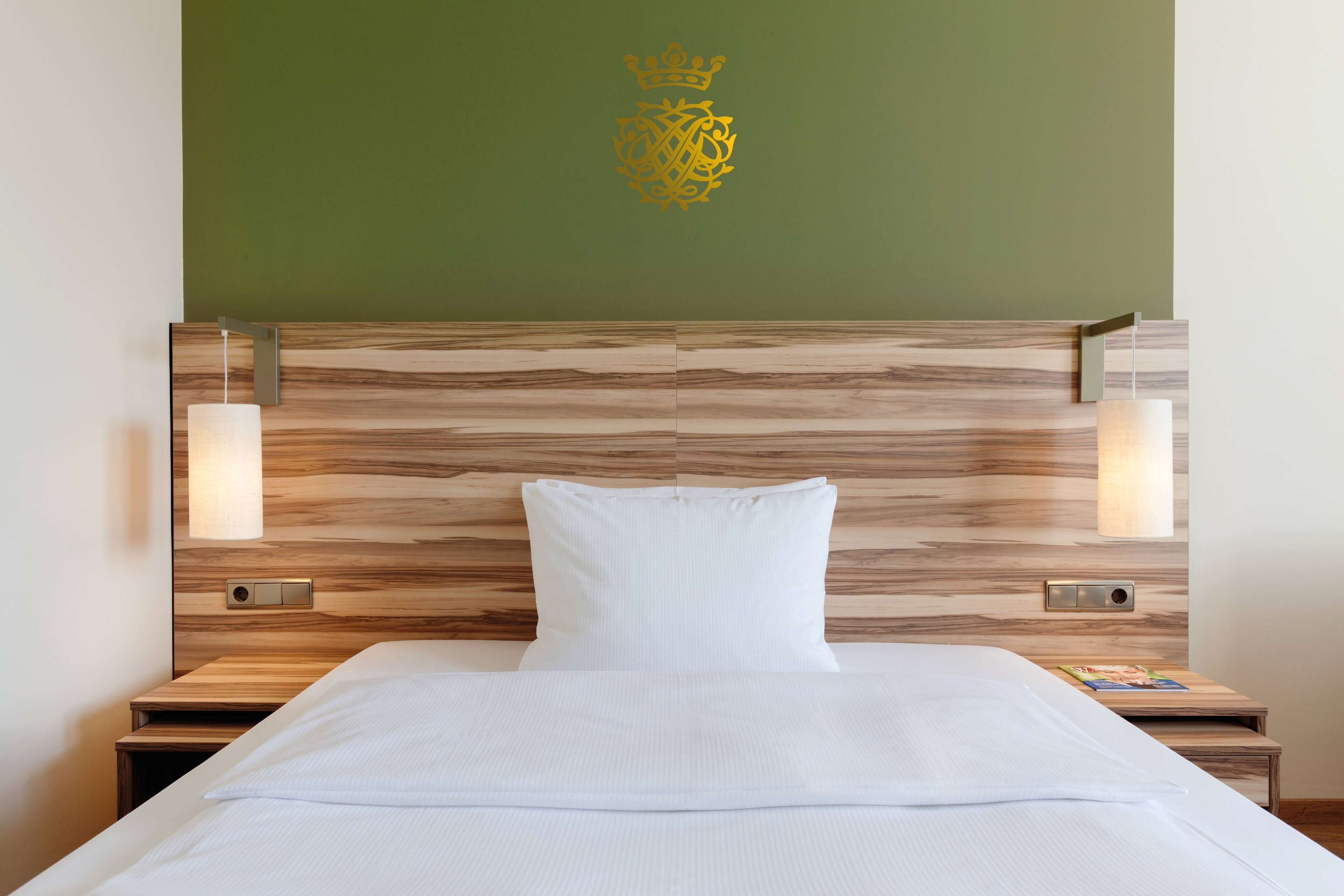 Un lit bien fait, avec du linge de lit blanc, une tête de lit en bois ornée d’un motif en forme de couronne, et deux lampes de chevet de chaque côté.