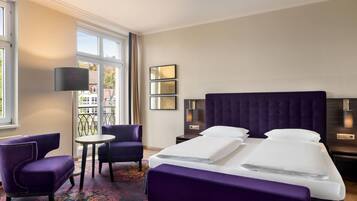 A hotel room with a large bed, two chairs, a small table, and a view of the outside.