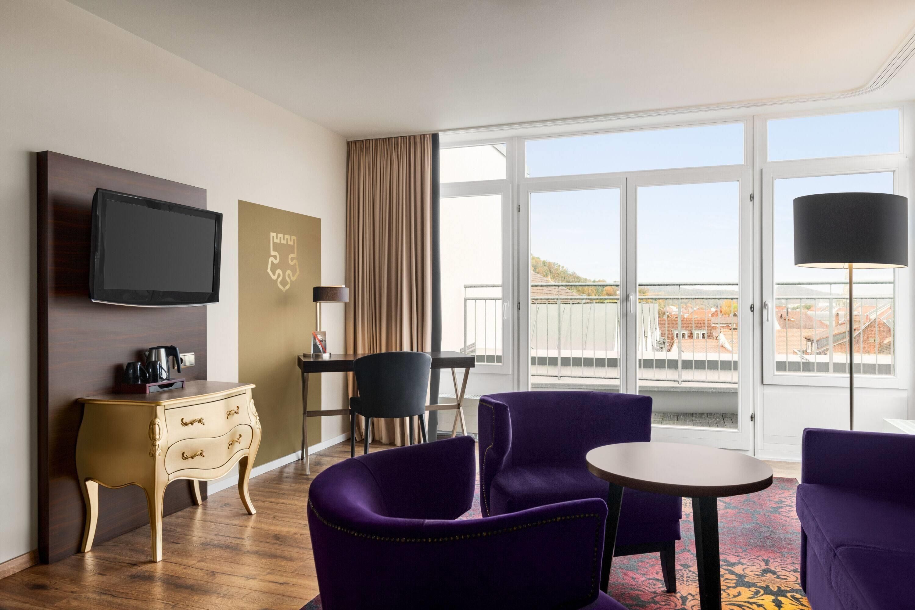 Ein Hotelzimmer mit Fernseher, einem kleinen Tisch, violetten Stühlen, einer Couch, einem Schminktisch und einem großen Fenster mit Aussicht.