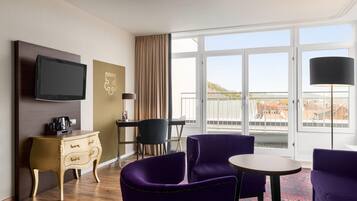 Ein Hotelzimmer mit Fernseher, einem kleinen Tisch, violetten Stühlen, einer Couch, einem Schminktisch und einem großen Fenster mit Aussicht.