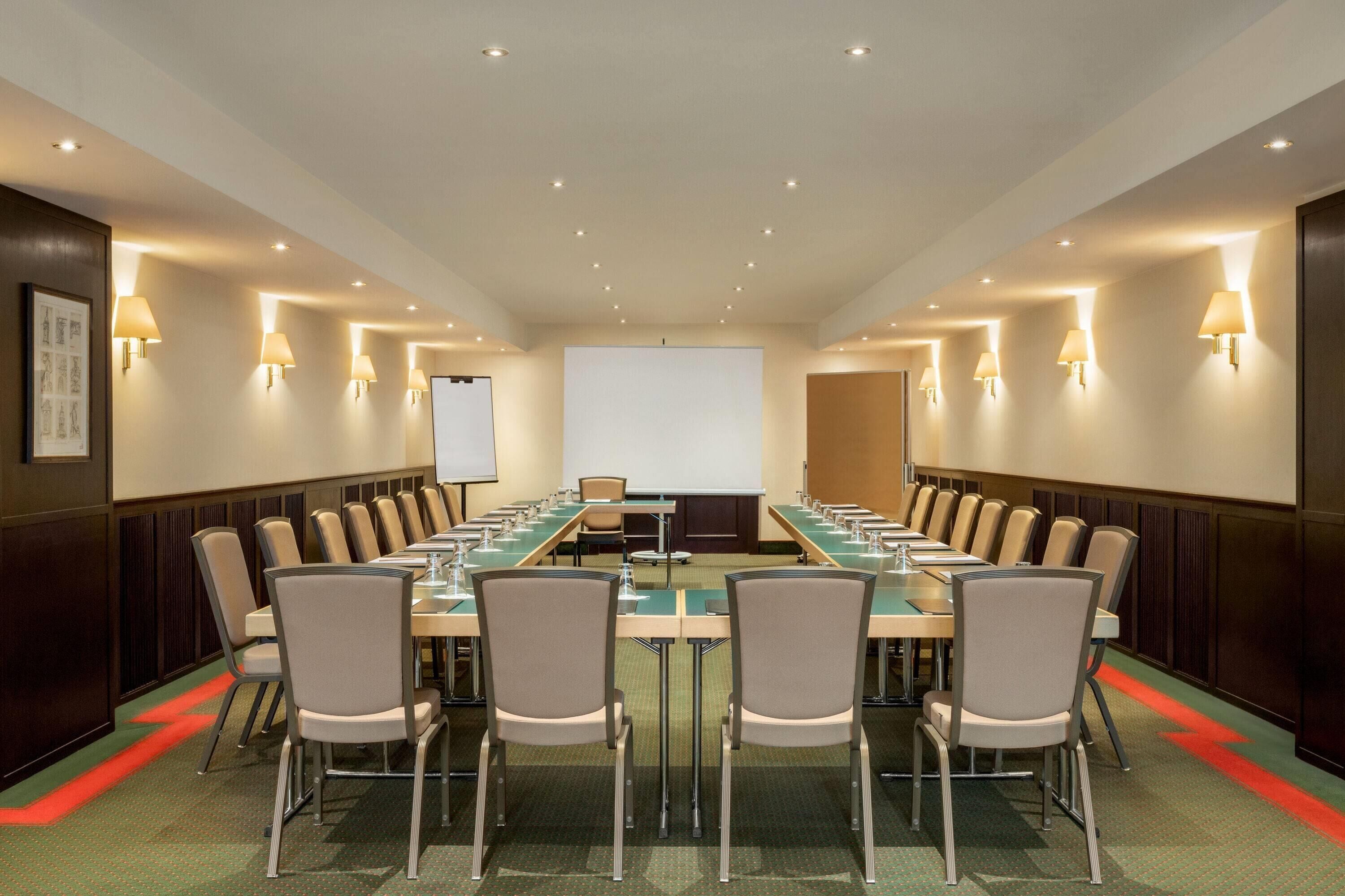 Uma sala de conferência com uma mesa comprida, cadeiras, tela de projetor e luminárias fixadas na parede.