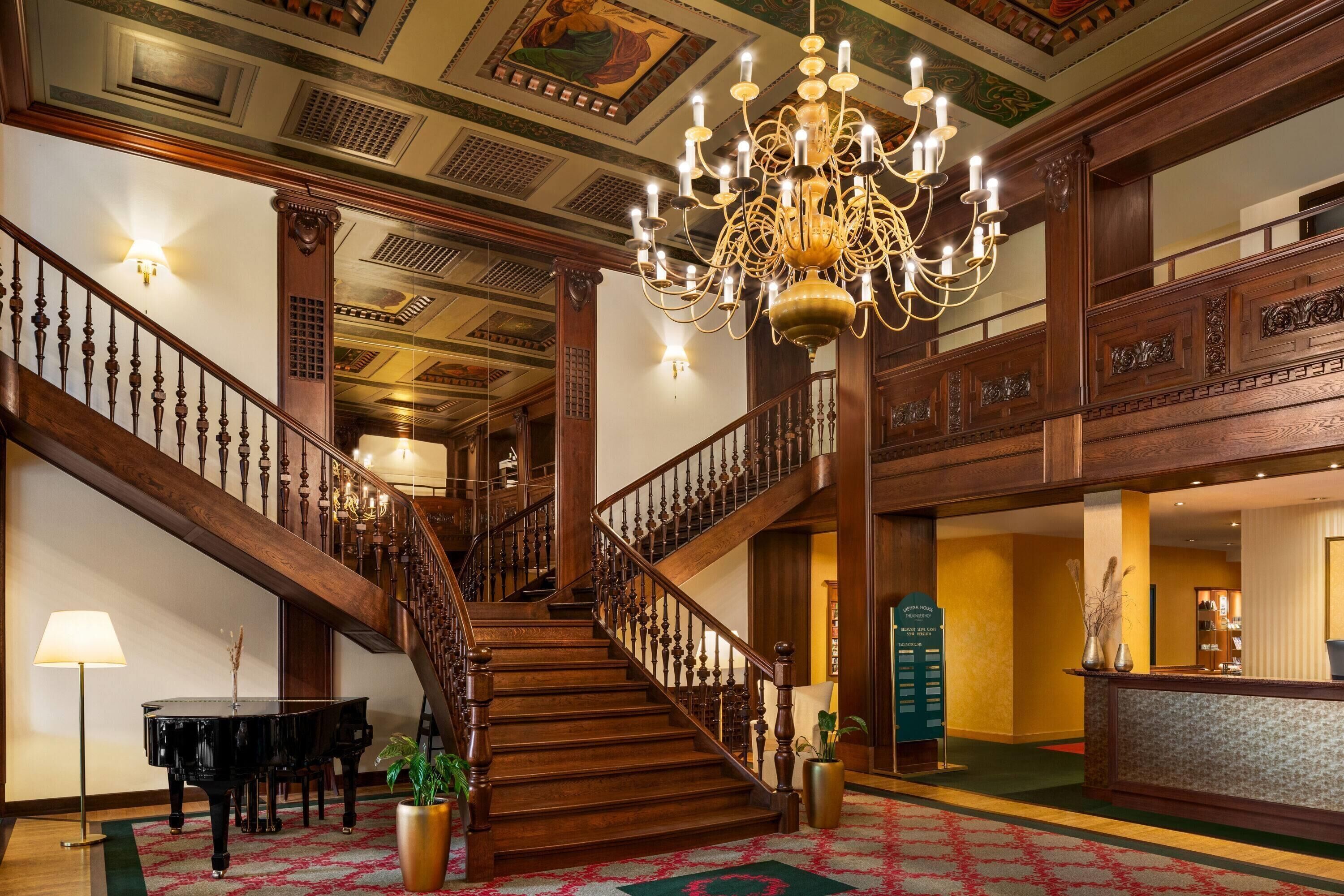 A grand staircase with wooden railings and a chandelier, a grand piano, and a reception desk in a hotel lobby.