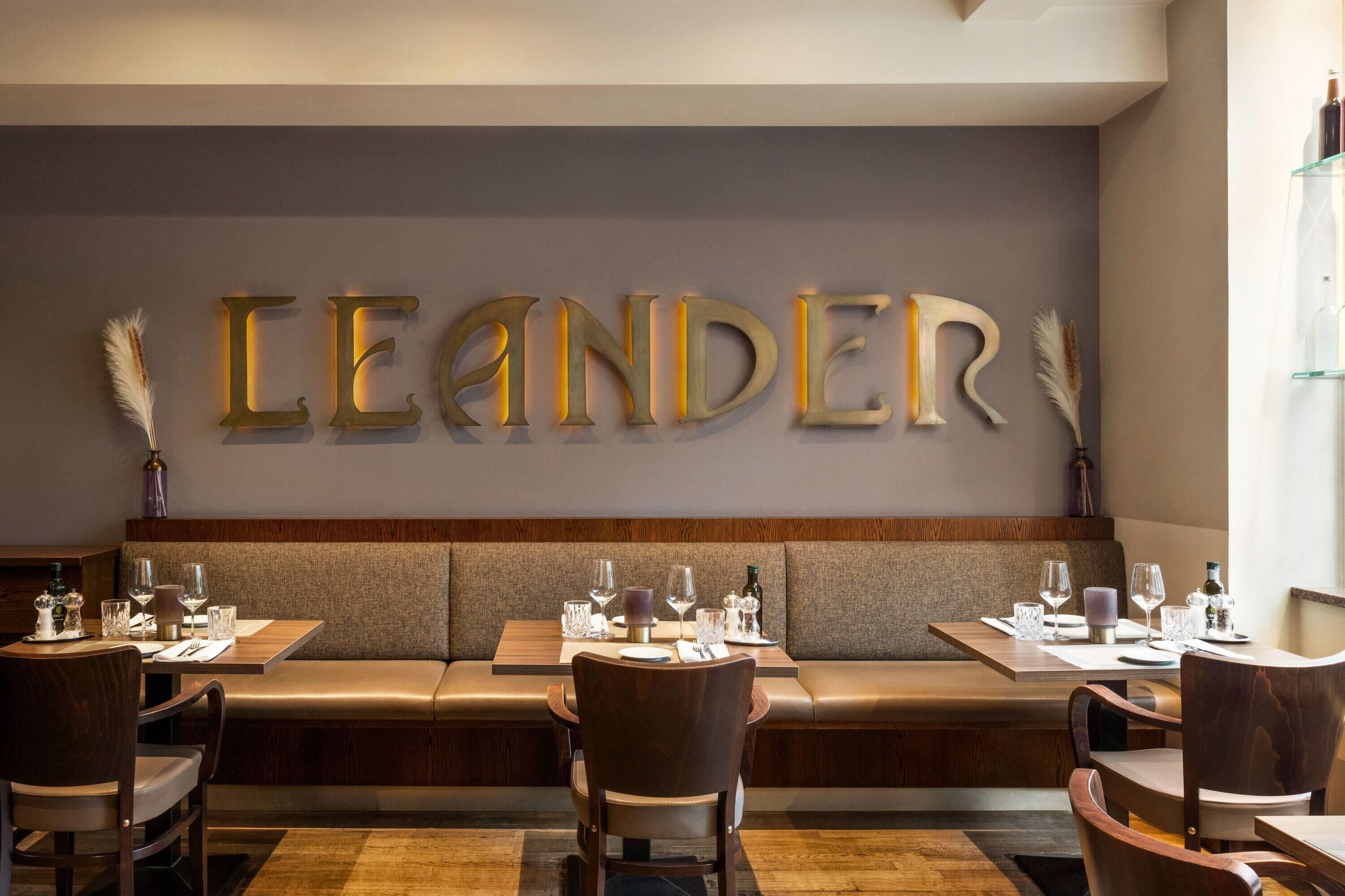 A restaurant interior with a long bench seating area, tables with chairs, and a wall with illuminated letters spelling 'LEANDER'.