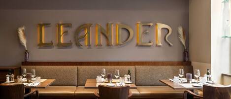 A restaurant interior with a long bench seating area, tables with chairs, and a wall with illuminated letters spelling 'LEANDER'.