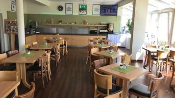 A restaurant with wooden tables and chairs, a kitchen area, and framed pictures on the wall.
