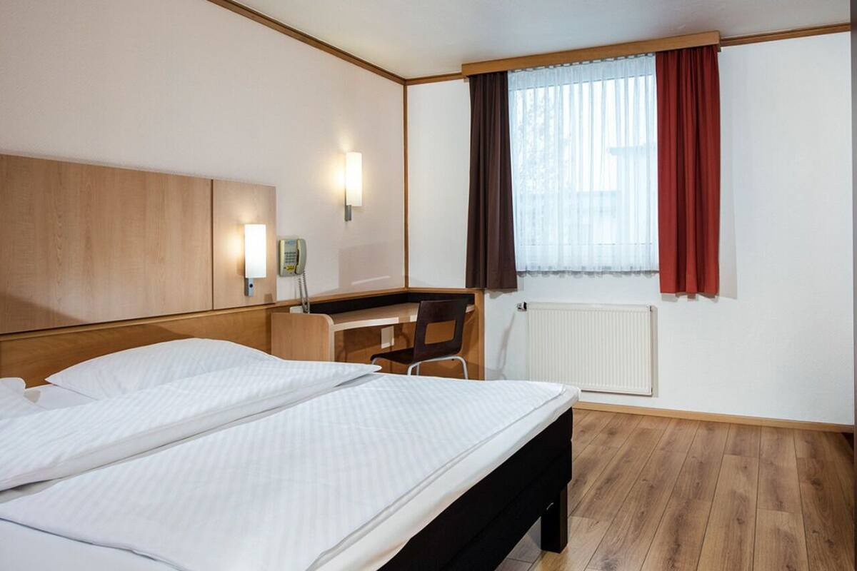 A hotel room with a large bed, a desk, a chair, a telephone, and a window with curtains.