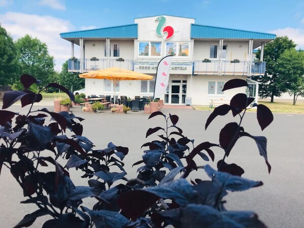 A hotel with a blue roof and a sign with a colorful logo. There is a covered outdoor seating area with tables and chairs. - Good Morning Erfurt (Erfurt)
