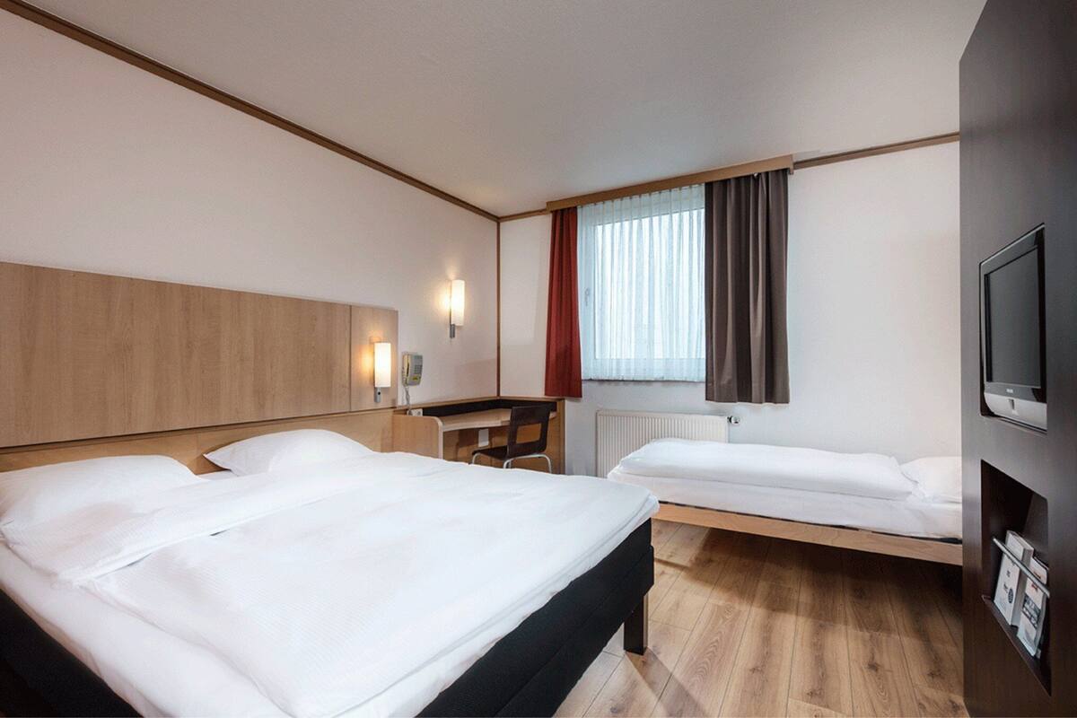 A hotel room with two beds, a desk, a chair, a TV, and a window with curtains.