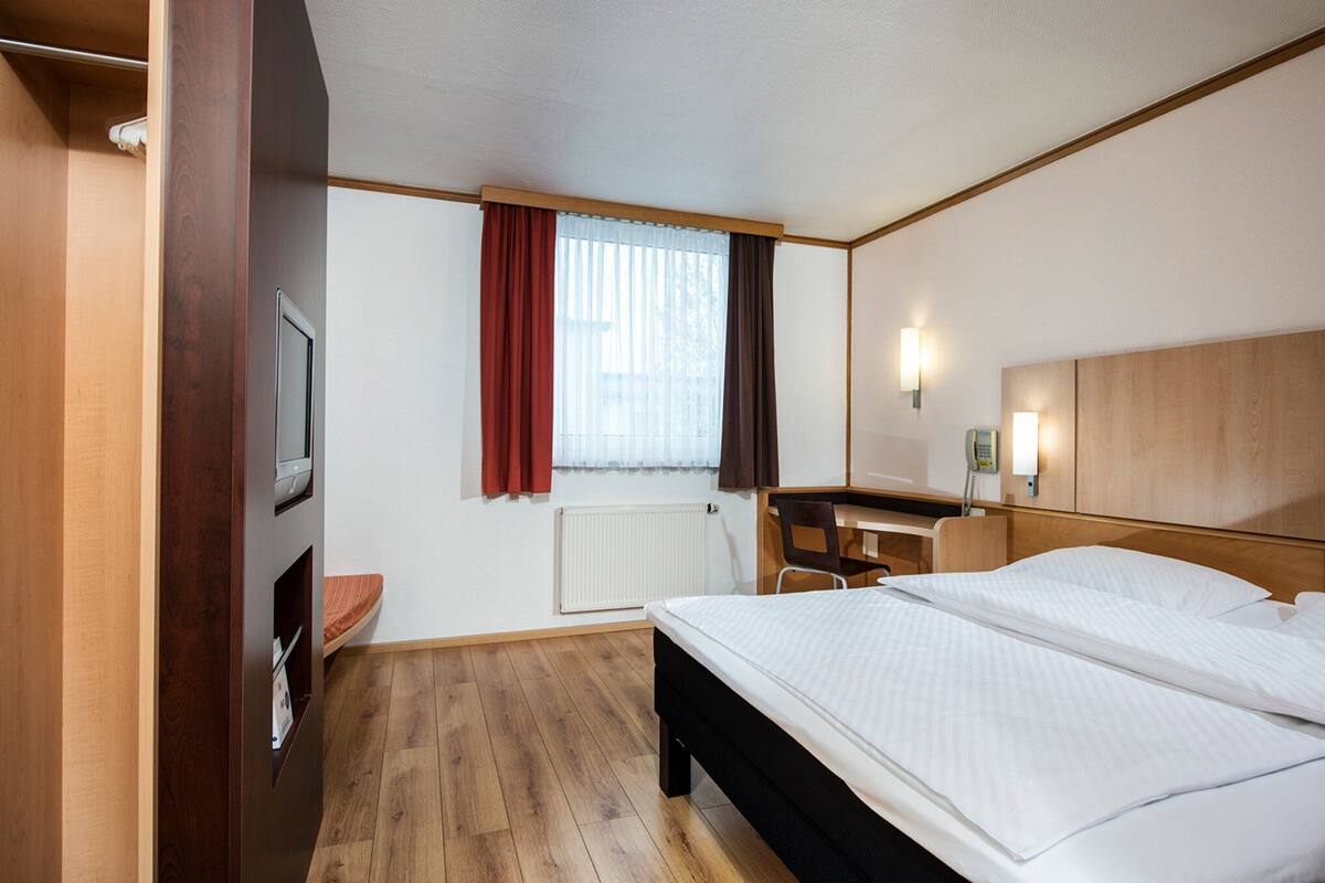 A hotel room with a bed, a desk, a chair, a TV, a window with curtains, and a wall-mounted light.
