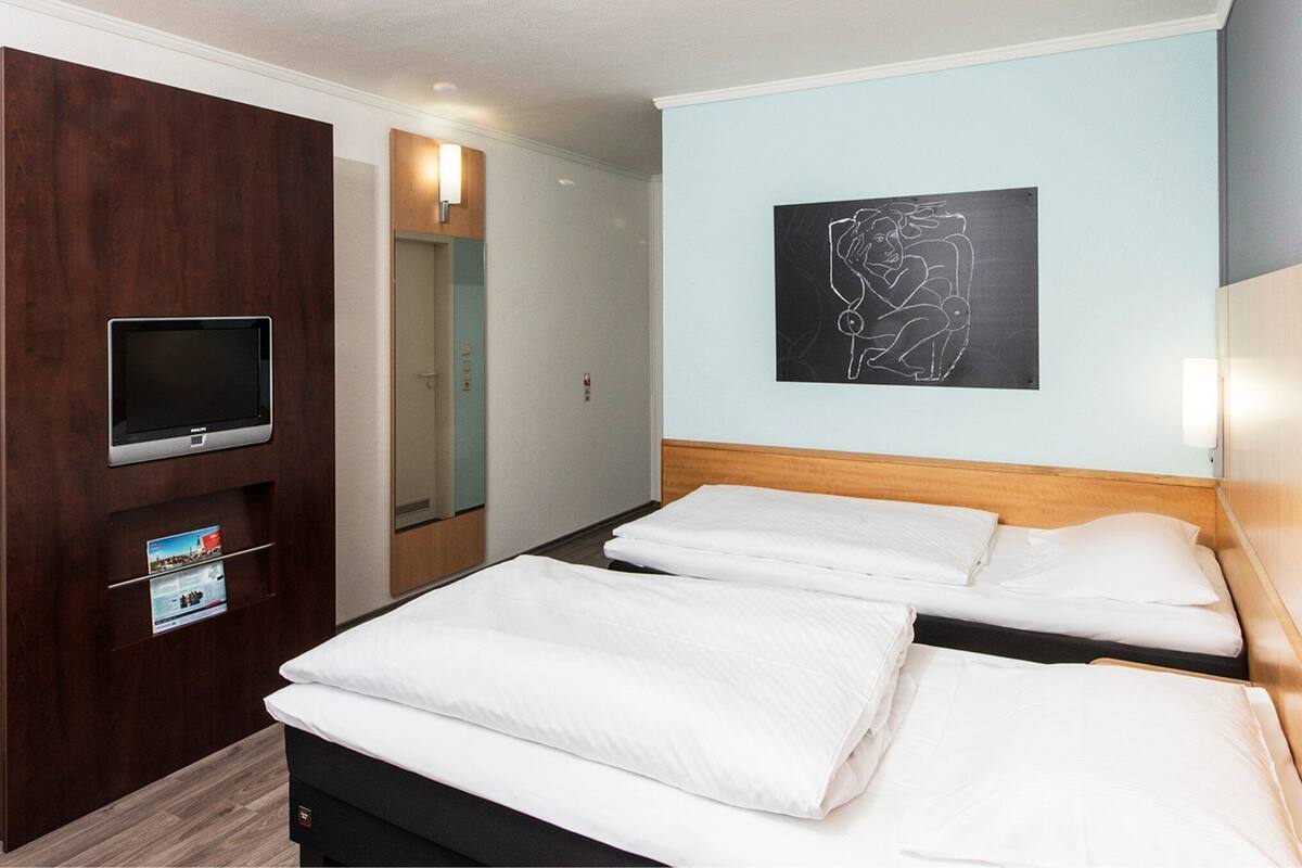 A hotel room with two beds, a TV, a wooden headboard, a wall art piece, and a mirror.