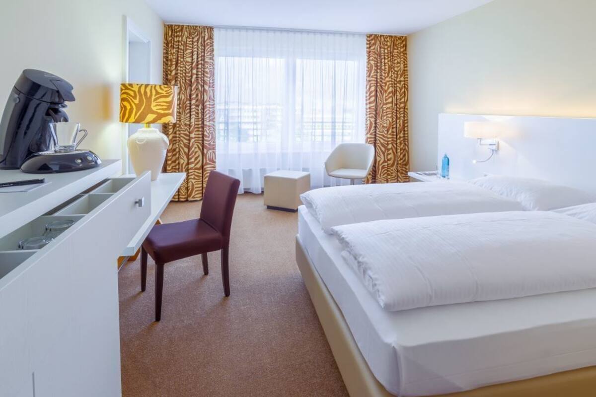 A hotel room with a bed, a desk, a chair, a coffee machine, and a lamp.