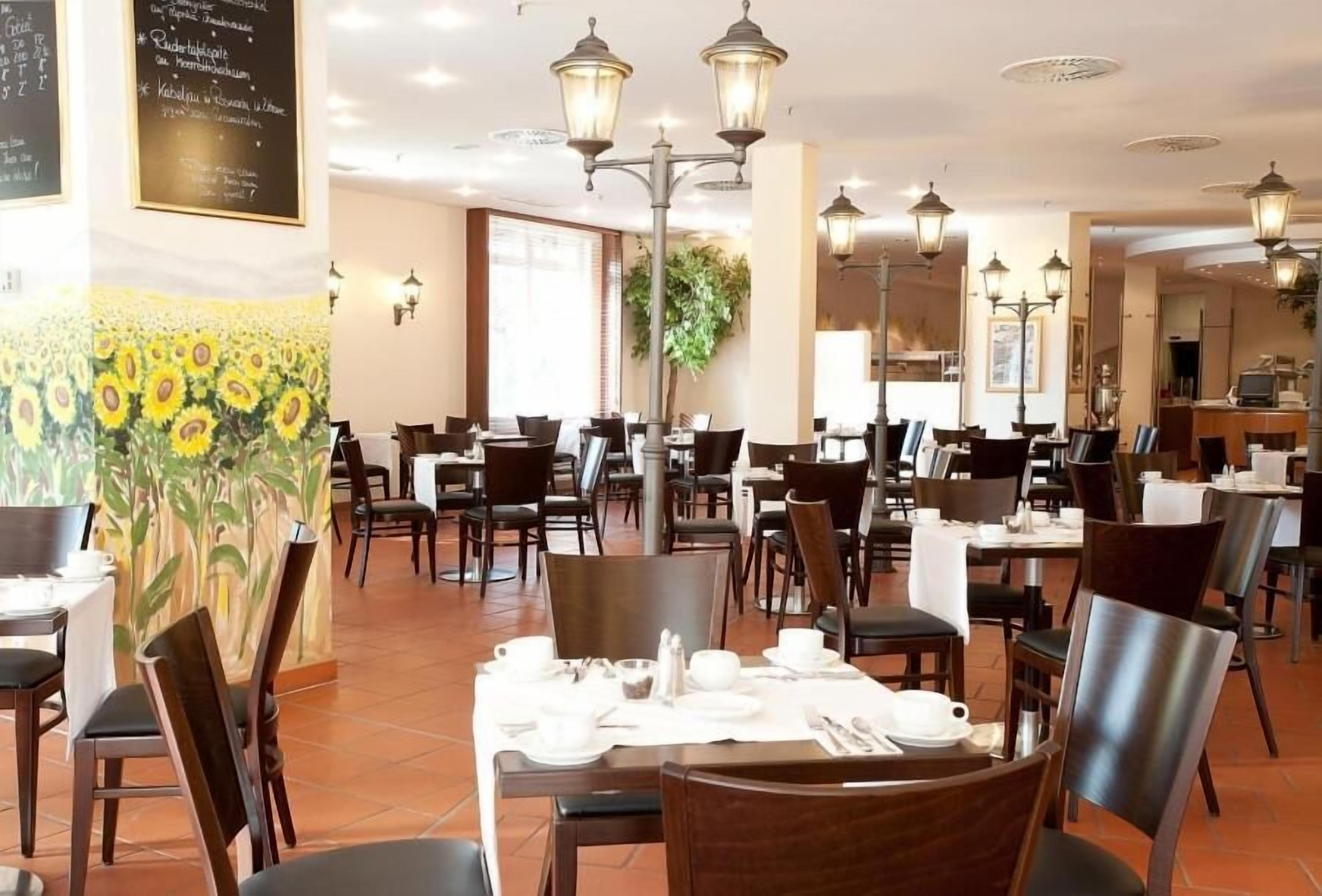 A restaurant with multiple tables and chairs, a mural of sunflowers, and hanging lantern-style lights.