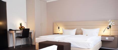 A hotel room with two beds, a desk with a chair, a wardrobe, and a bedside table with a lamp and flowers.