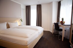 A hotel room with a double bed, a desk with a chair, and a fruit bowl on the desk. - Manhattan Hotel (Frankfurt)