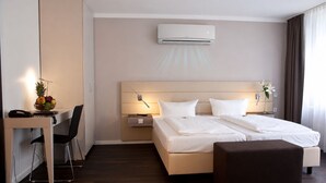A hotel room with a bed, bedside tables, a desk, a chair, an air conditioner, and a window with curtains. - Manhattan Hotel (Frankfurt)