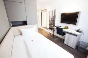 A modern hotel room with a bed, a desk with a chair, a TV, and a small trash bin. - Manhattan Hotel (Frankfurt)